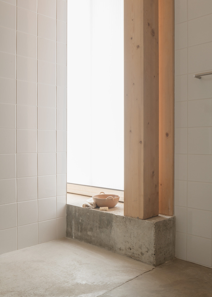 White tiled bathroom corner with tall window and concrete sill holding a wooden bowl