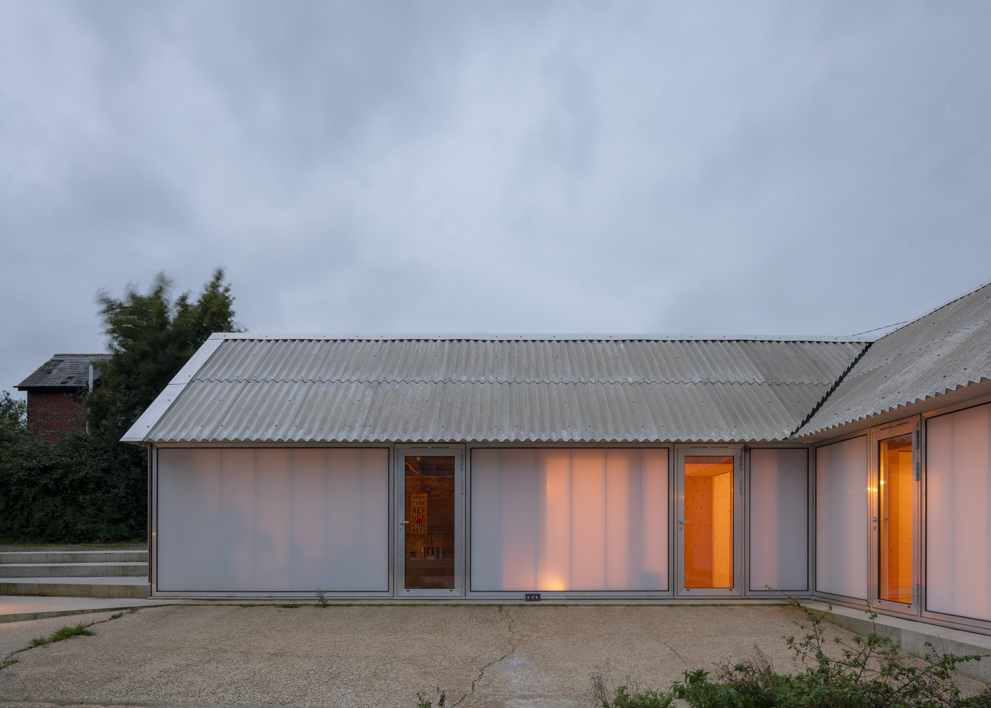 Corrugated metal roof over translucent facade panels glowing at dusk beside gravel courtyard