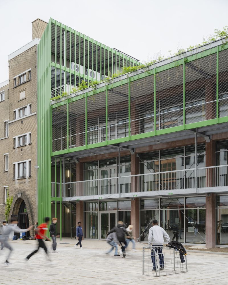 Street view of the green steel and glass facade with pedestrians crossing the paved courtyard below
