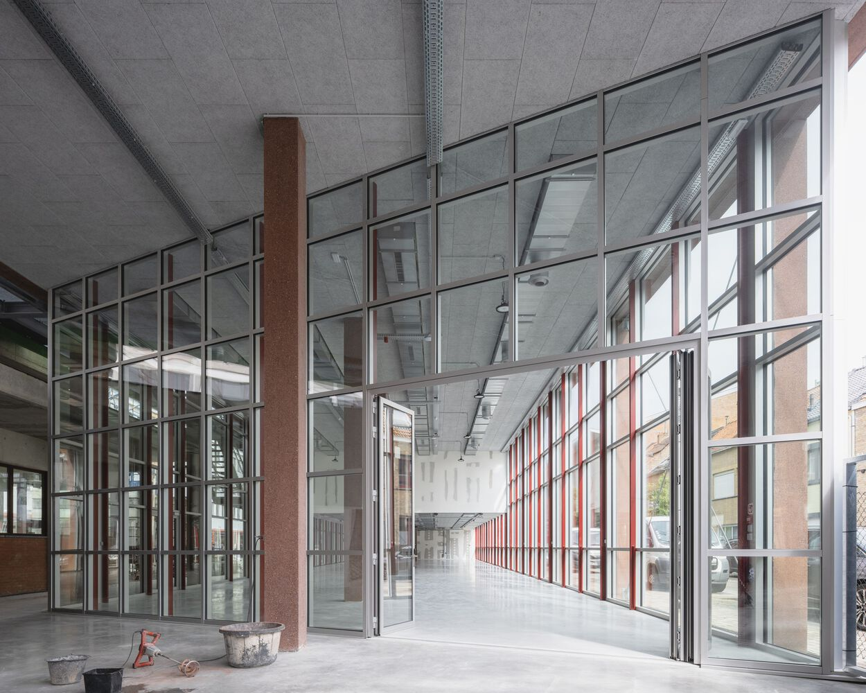 Interior corridor with floor-to-ceiling glass partitions and red steel mullions under a concrete ceiling
