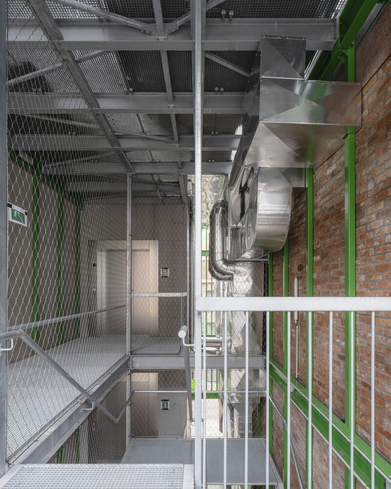 Stairwell with wire mesh enclosures, exposed ductwork, and green steel framing along brick wall