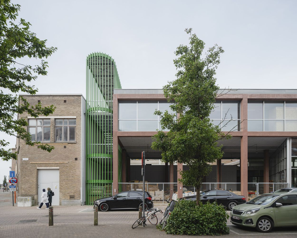 Street view with the green vertical fin tower next to brick buildings and parked cars under overcast sky