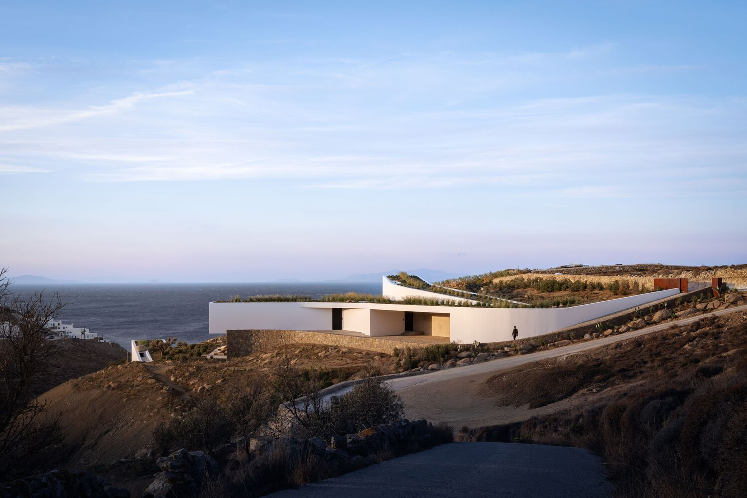 Distant view of the white volumes and cantilevered roof nestled into the rocky coastal hillside at dusk