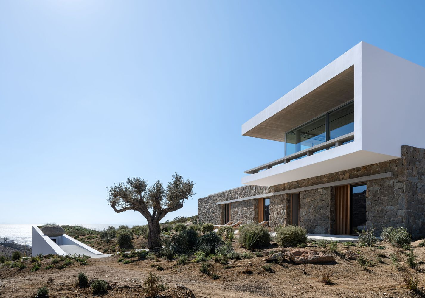 Stone-clad lower volume with timber openings beneath the white cantilevered upper floor and an olive tree