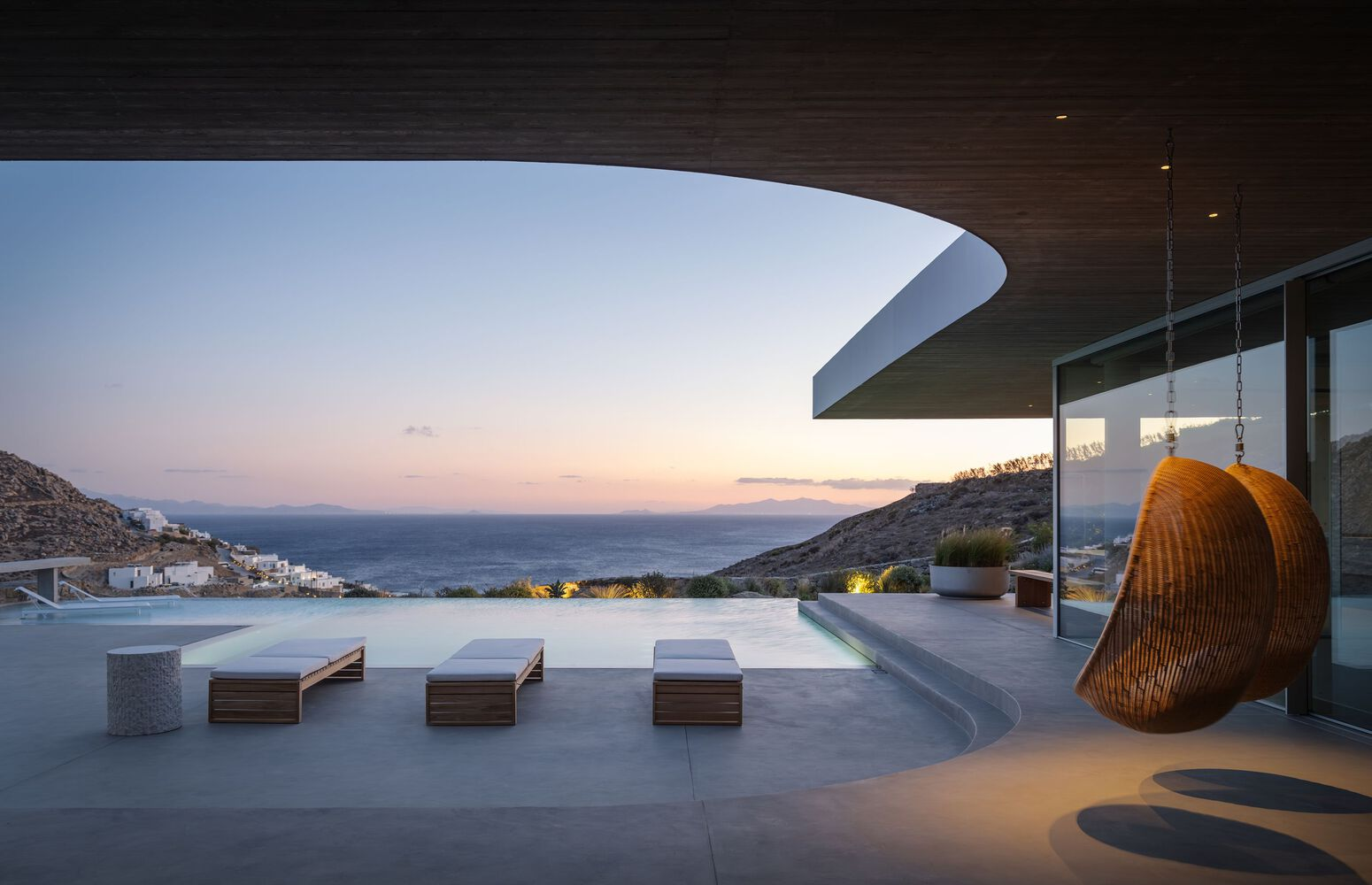 Poolside terrace with woven hanging chair and curved roof overhang at dusk