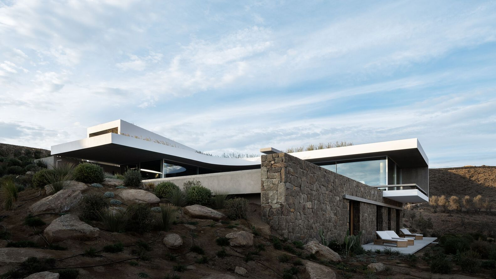 Stacked volumes with stone retaining walls and cantilevered roof planes amid boulders and native grasses