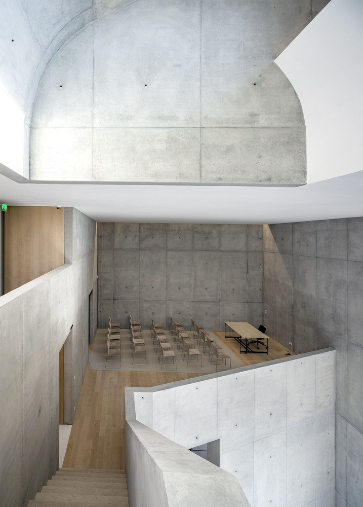 Curving concrete auditorium with board-formed walls and rows of timber seating below vaulted ceiling