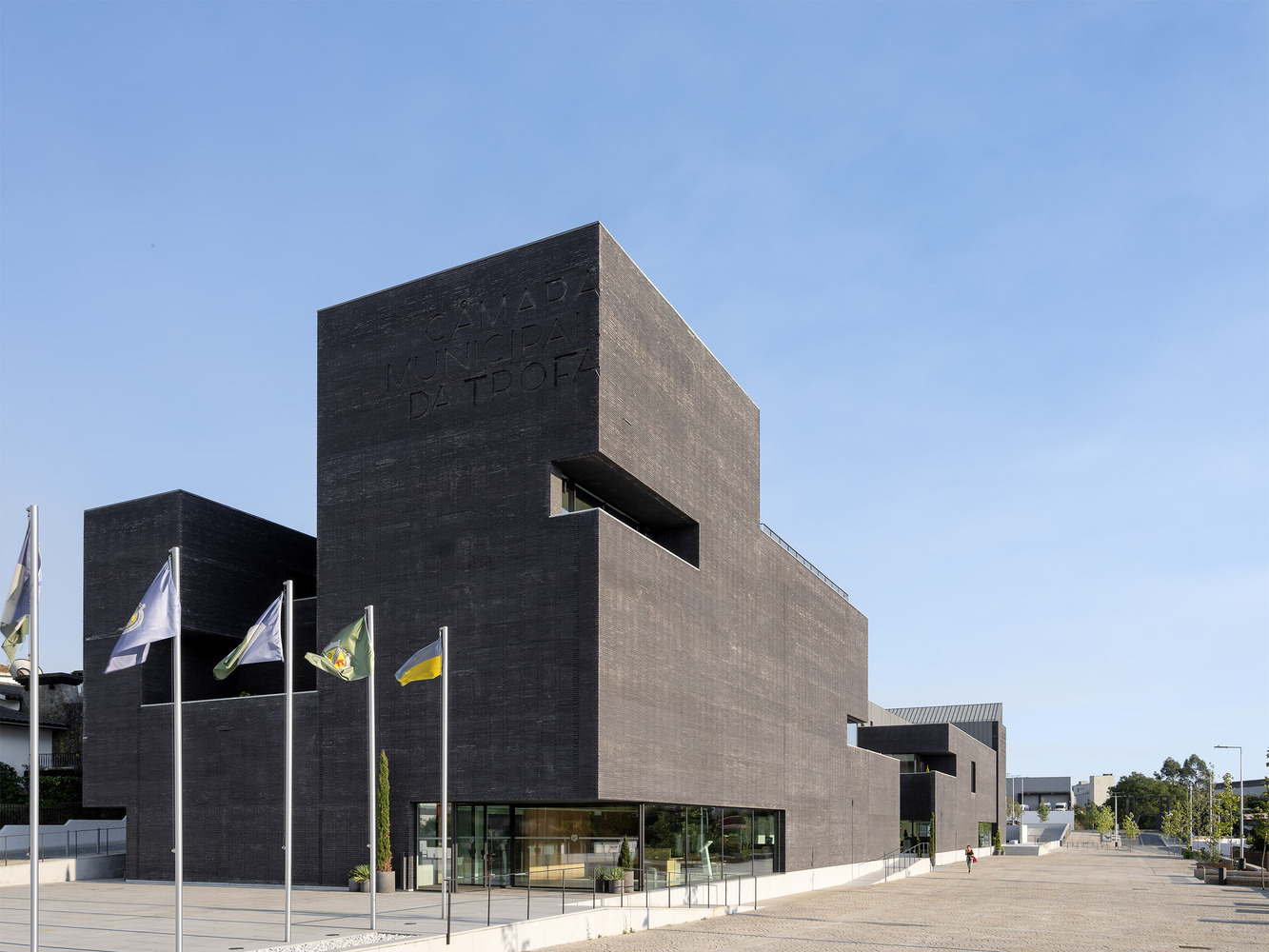 Dark brick volume with horizontal window openings and flagpoles along the plaza under clear blue sky
