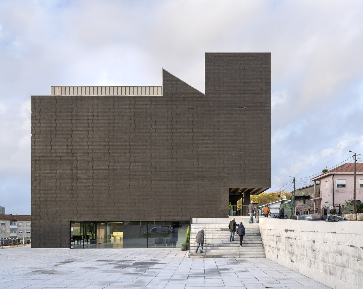 Rear facade of dark brick volumes with concrete stairs ascending to the entrance in overcast weather