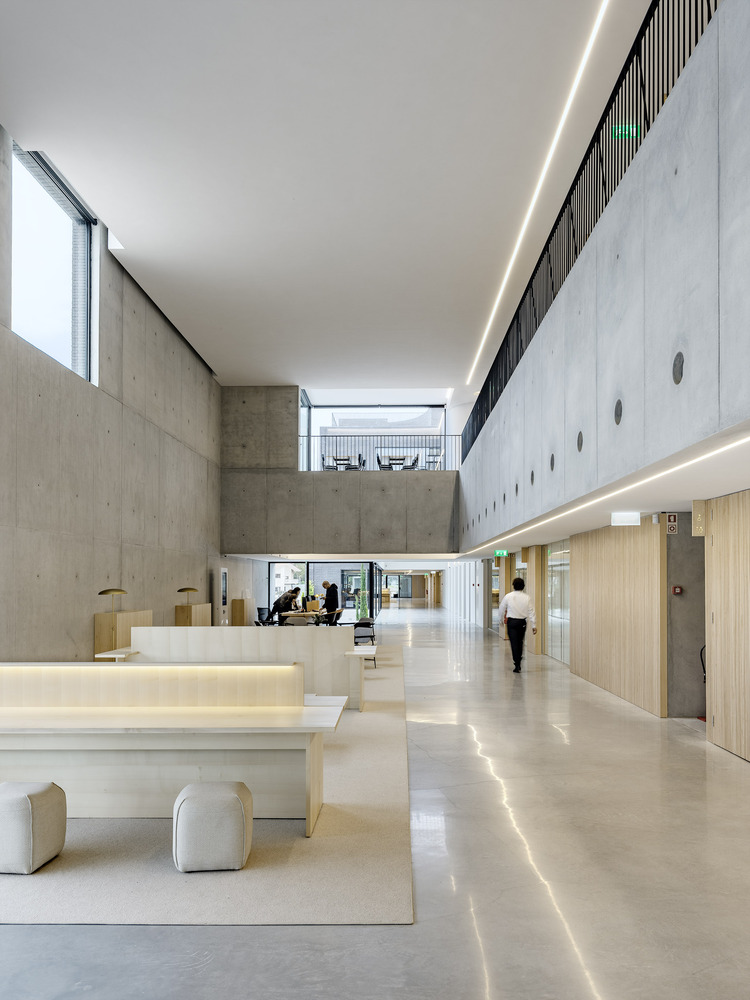 Double-height lobby with exposed concrete walls, wooden reception desk, and a person walking through diffuse daylight