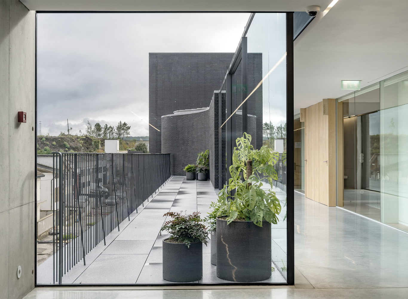 View through glass doors to rooftop terrace with dark brick volumes and planters under cloudy sky