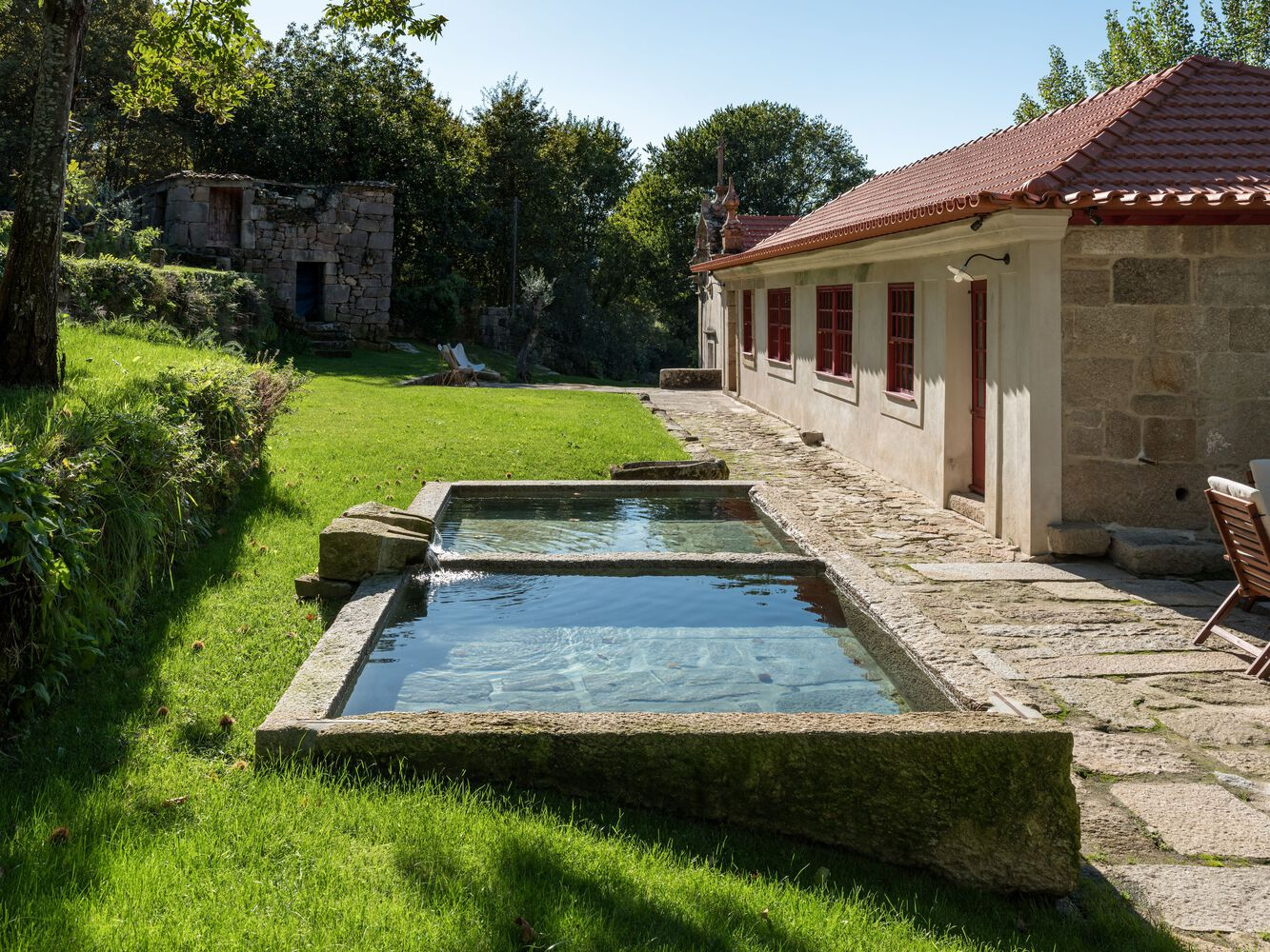 Stone-edged reflecting pool adjacent to a single-story stone residence with red tiled roof under trees