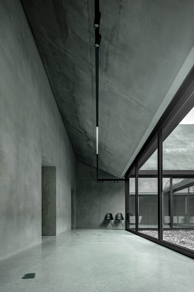 Interior room with angled concrete ceiling, polished floor, and glazed courtyard wall with two chairs