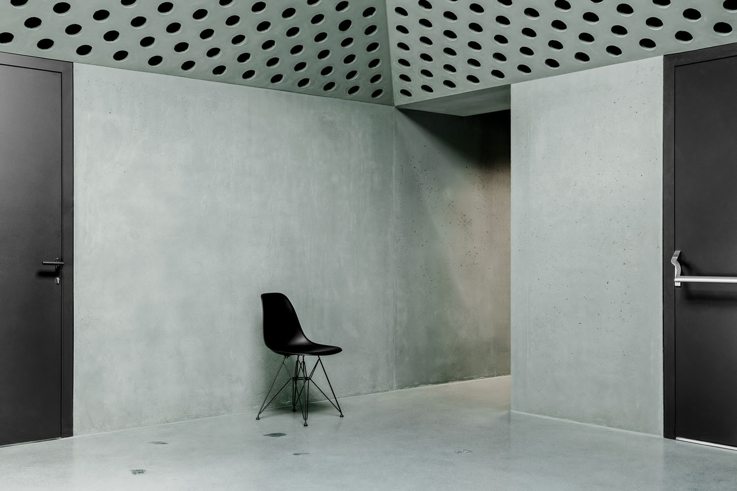 Corner of room with perforated ceiling, polished concrete walls, dark doors, and wire-frame chair