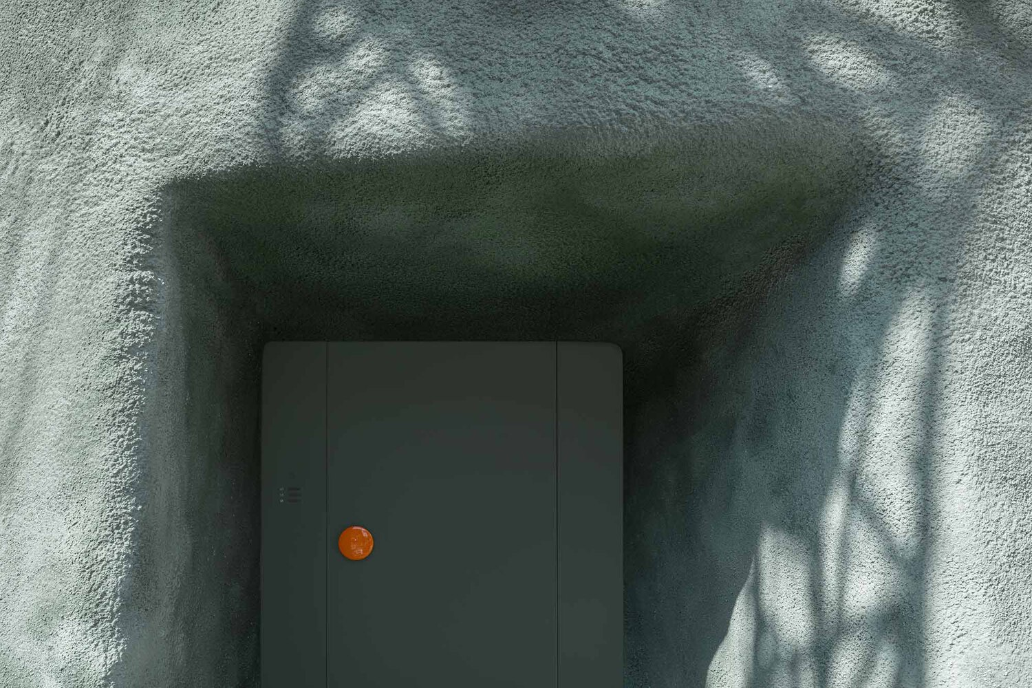 Textured stucco corridor with dappled shadows leading to grey door with orange circular handle