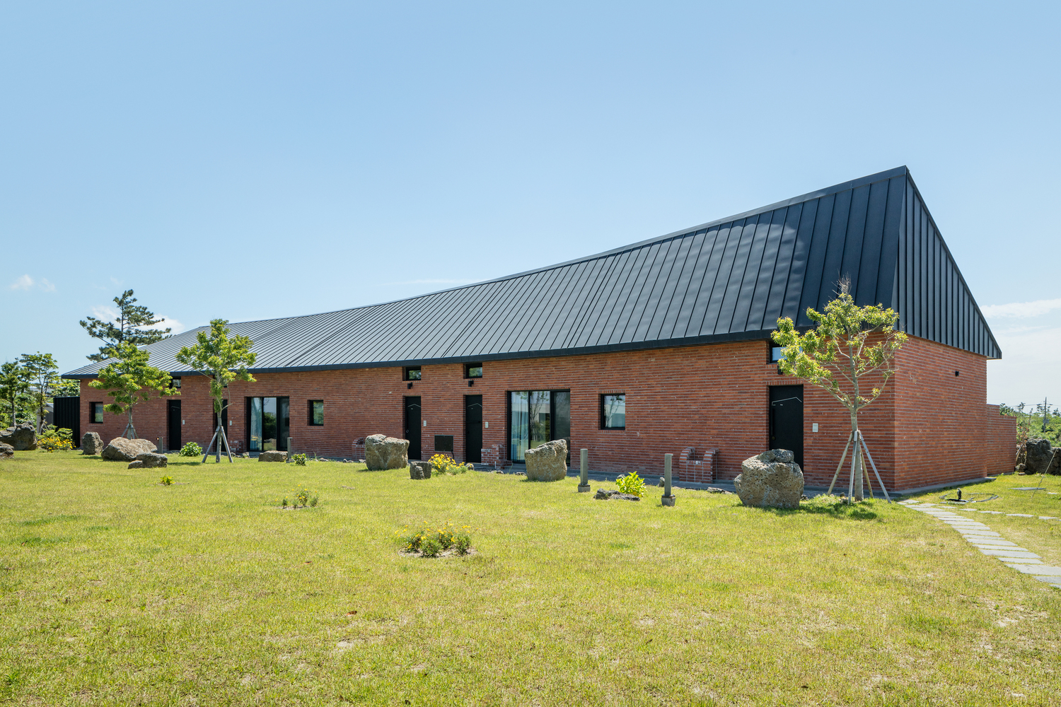Red brick base with charcoal metal roof volume set in a grass field with scattered boulders