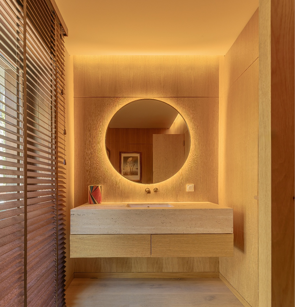 Bathroom vanity in terrazzo with circular backlit mirror and wood louver shutters filtering evening light