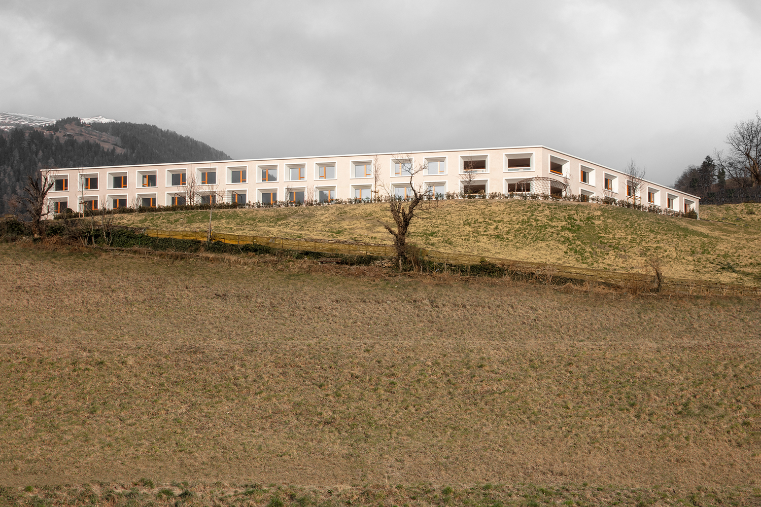 Long white facade with rhythmic window openings set on a grassy slope under an overcast sky