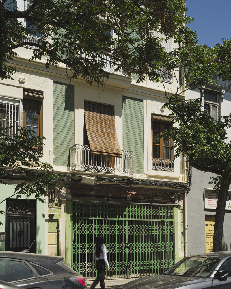 Street facade with green shutters, balconies with ornate ironwork, and a pedestrian passing below a tree