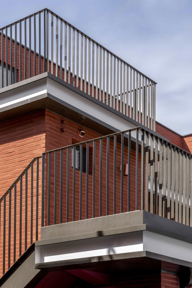 Stacked balconies with vertical metal railings and red brick cladding under a cloudy sky