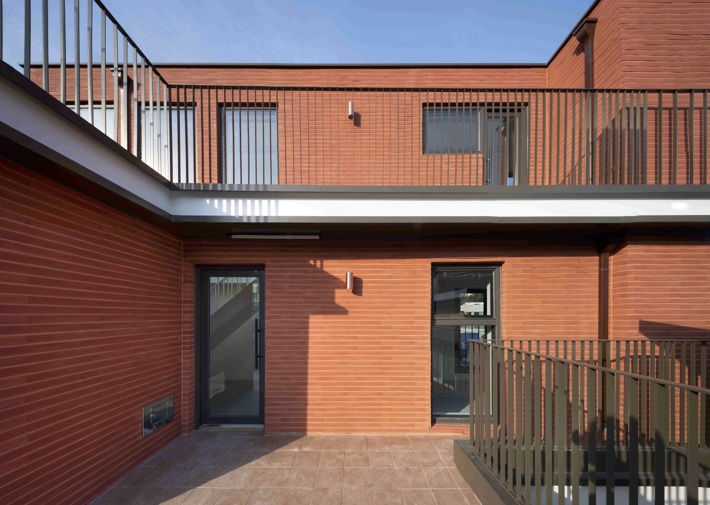 Courtyard with red tile facades, black entry doors, and metal railings under afternoon sun
