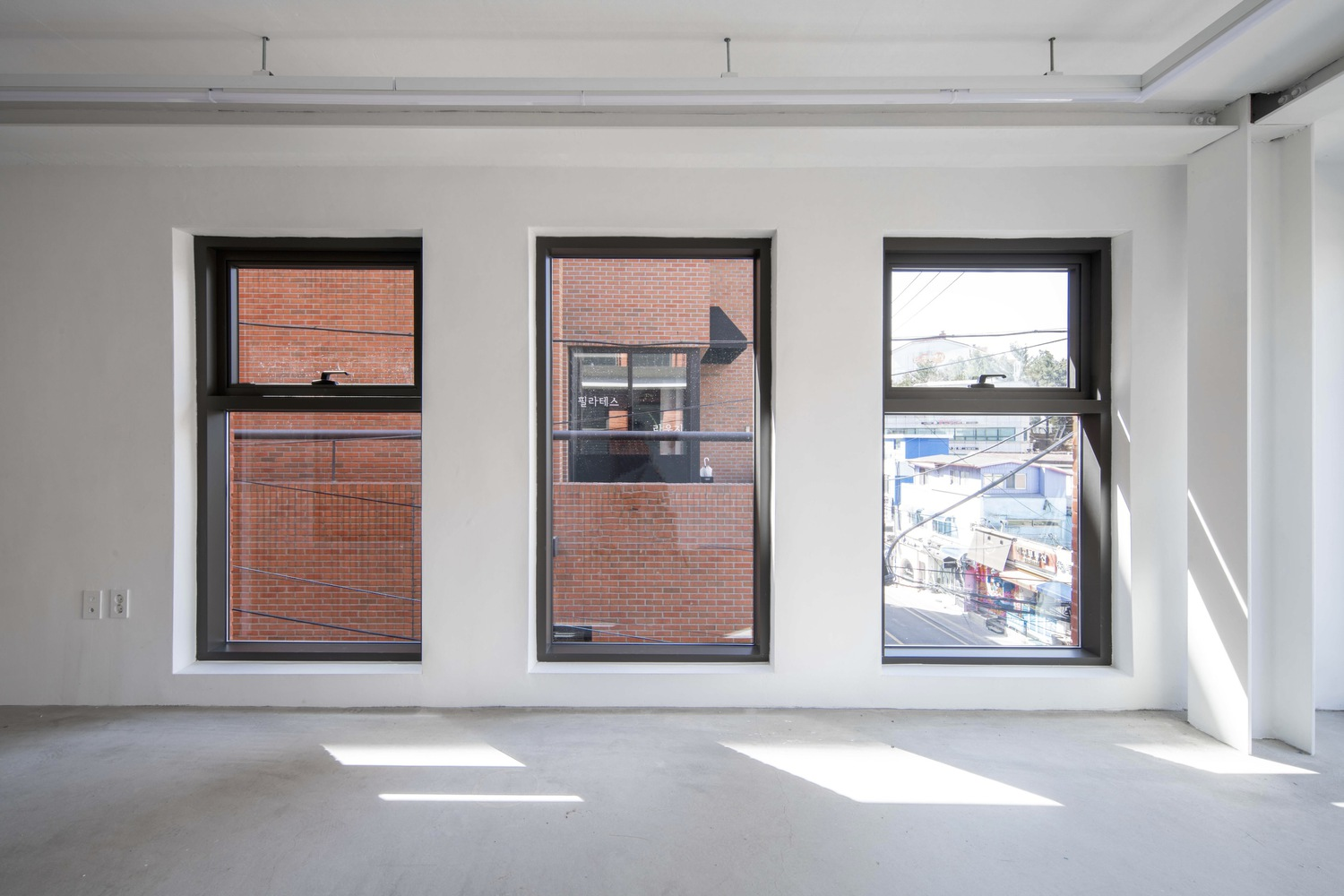 Interior view of three black-framed windows overlooking brick buildings with sunlight casting shadows on the concrete floor