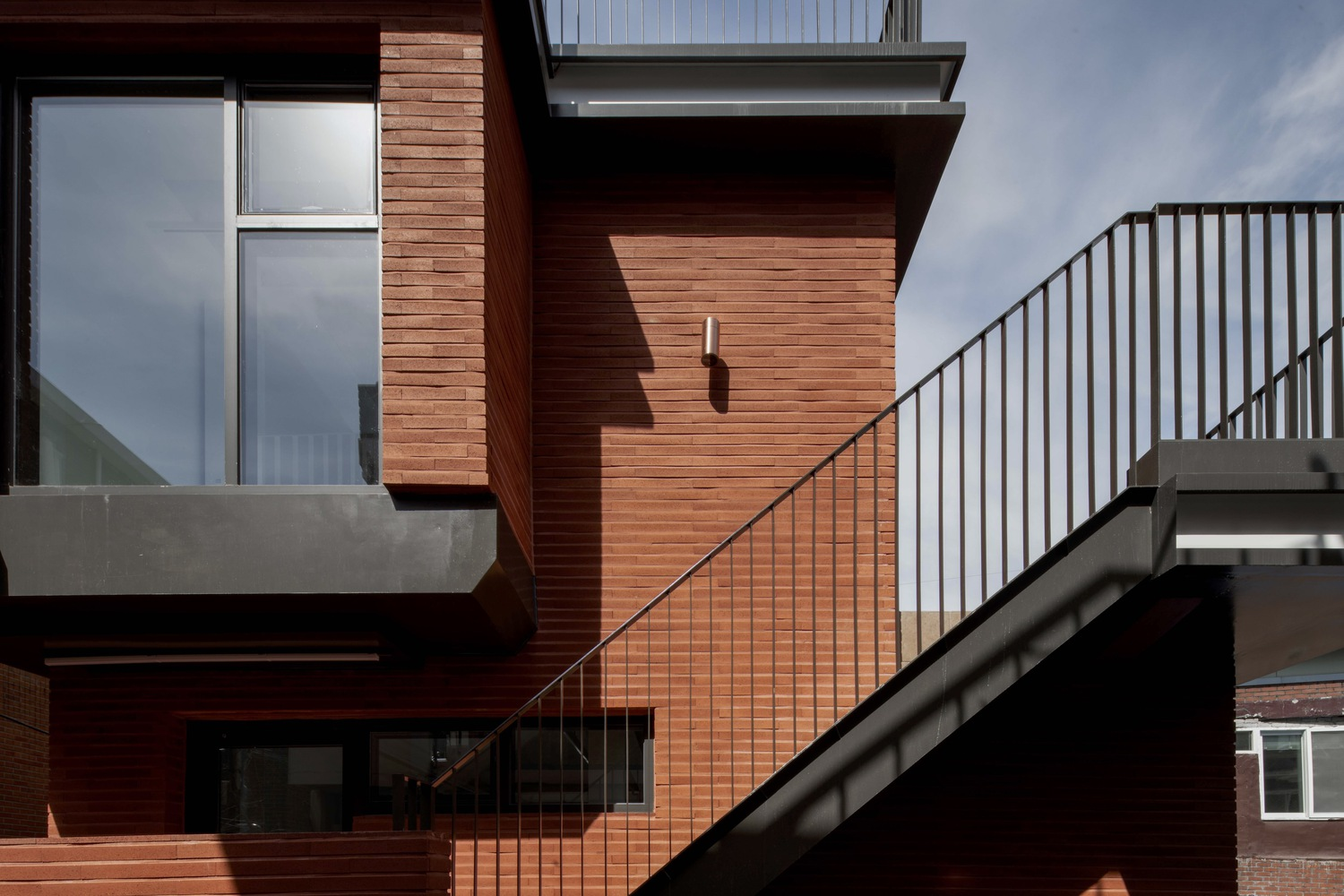Exterior stair with metal railing ascending between red brick clad volumes under afternoon sun