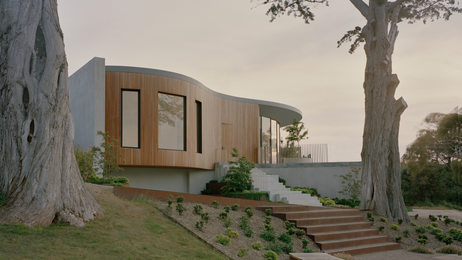 Curved timber-clad volume elevated on a concrete base with exterior stairs ascending through landscaped garden terraces