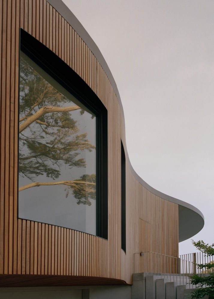 Close-up of the vertical timber cladding wrapping the curved facade with an angled window reflecting nearby trees