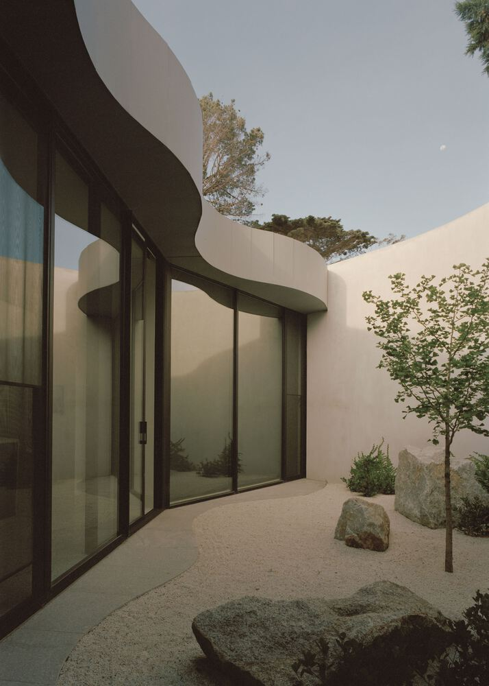 Courtyard with curved glazed walls and gravel surface planted with boulders and a young tree under dusk sky