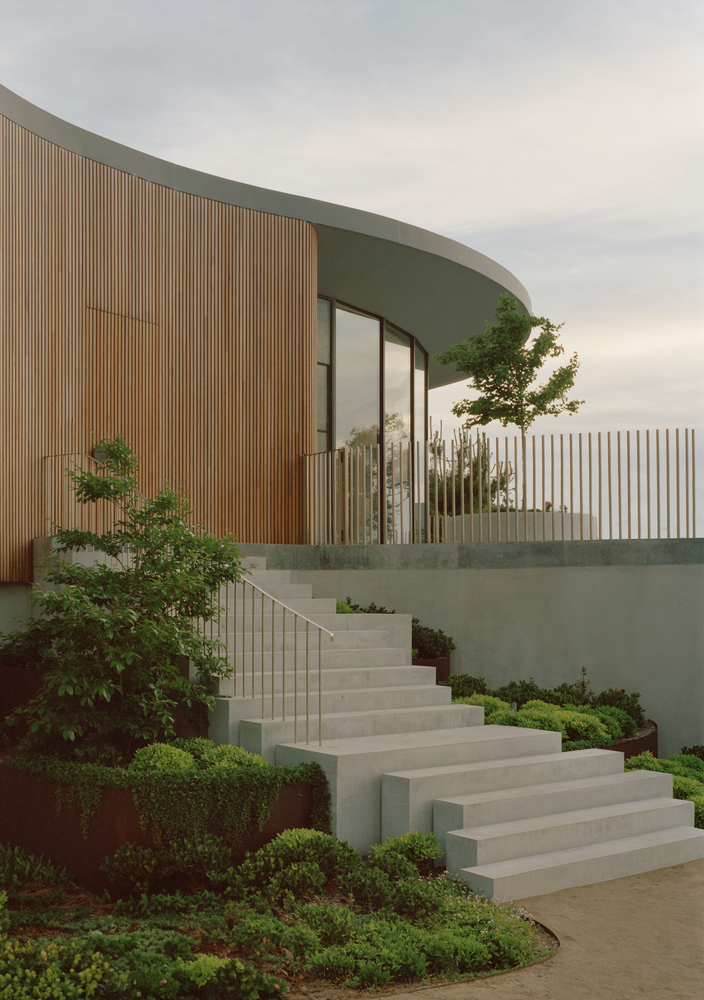 Concrete stairs with planted beds climbing toward the timber facade and metal balustrade under overcast evening sky