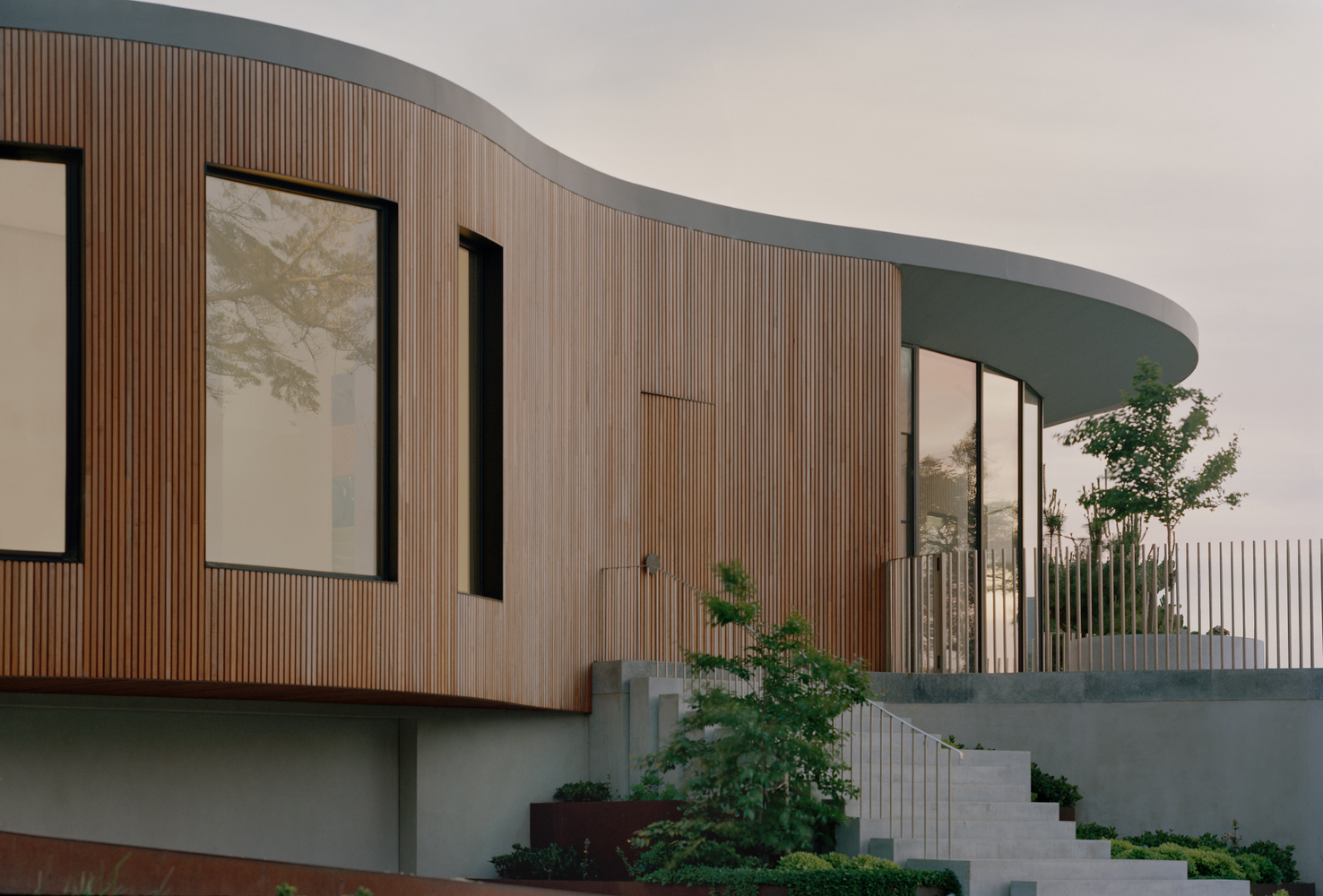 Curved timber-clad facade with vertical slats and dark-framed windows above a planted terrace