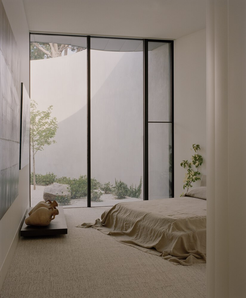 Minimalist bedroom with floor-to-ceiling glazing framing a white courtyard wall and sparse plantings