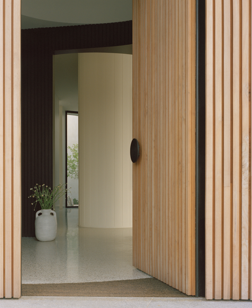 Vertical timber slat door pivoting open to reveal a curved plaster wall and ceramic vase