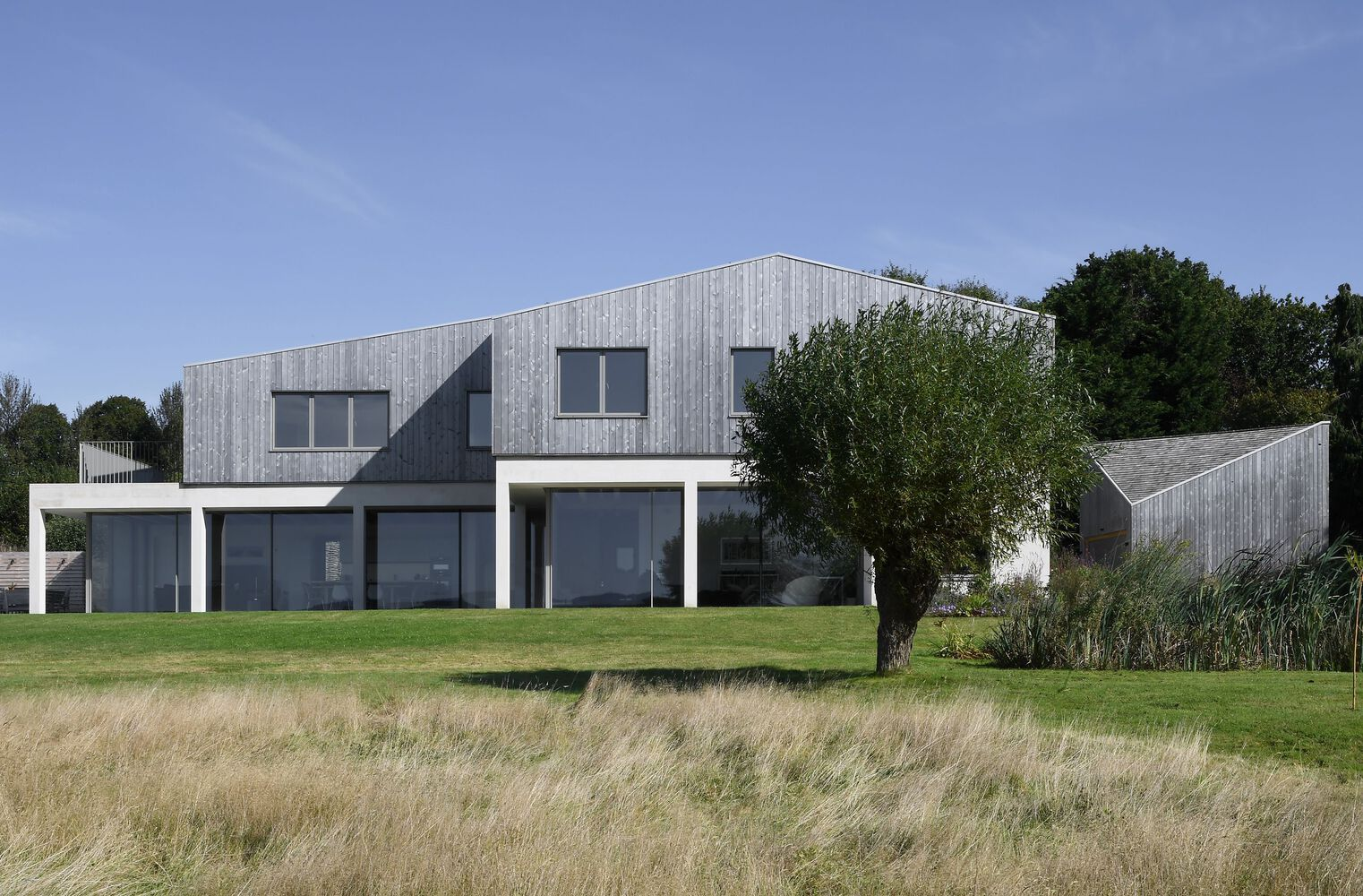 Garden facade with vertical timber cladding and white render base seen across dry grass meadow