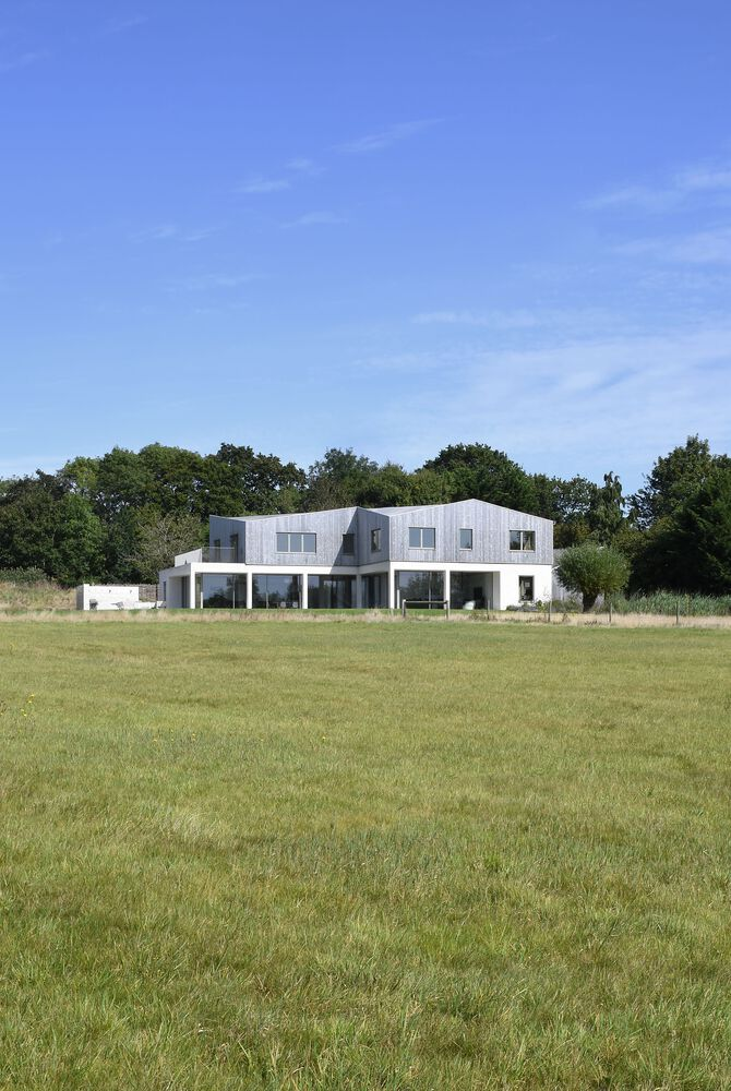 Rear elevation showing white rendered facade with upper shingle cladding across a mown meadow under blue sky
