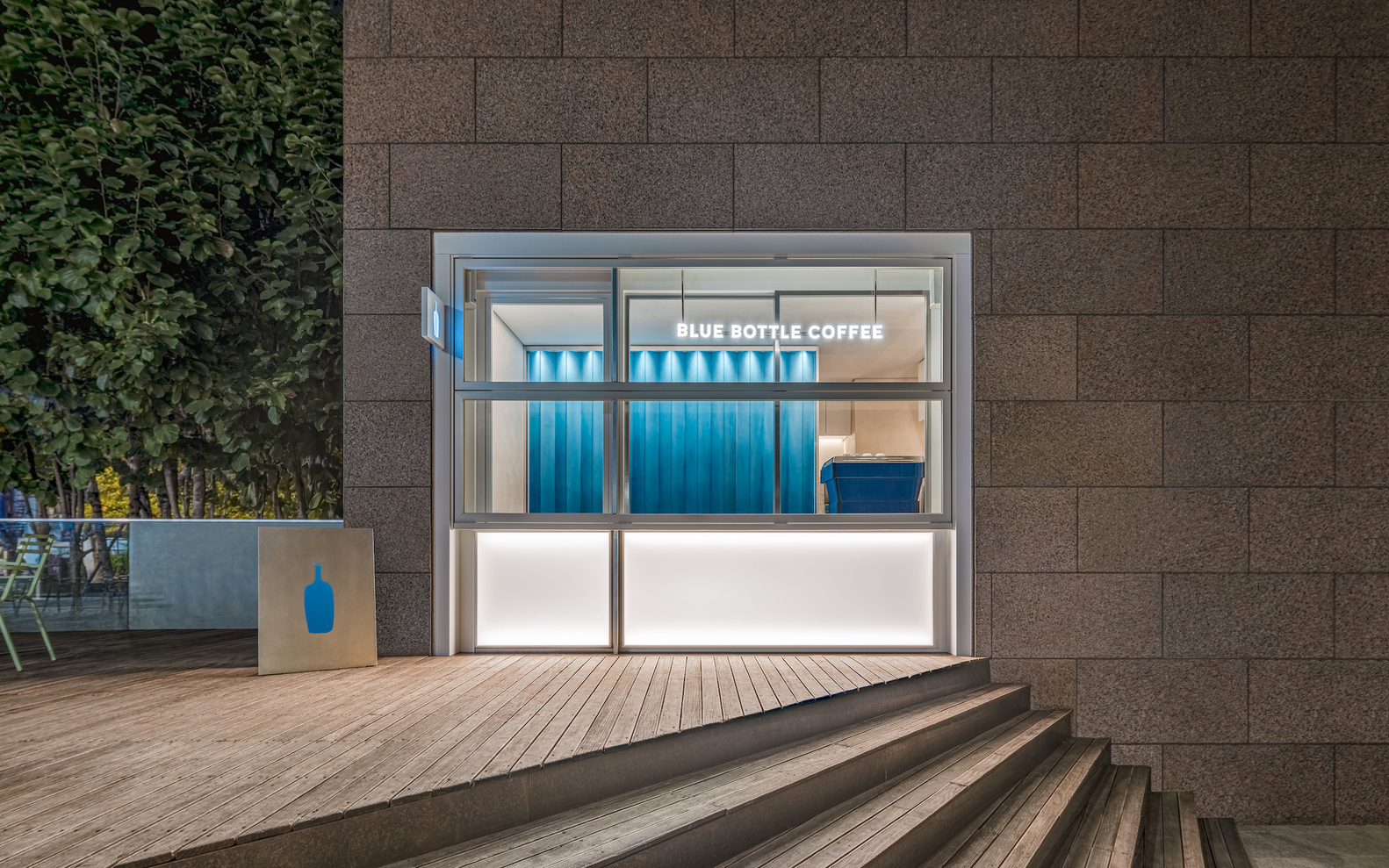 Glass-fronted kiosk with blue corrugated metal wall and timber deck steps at dusk