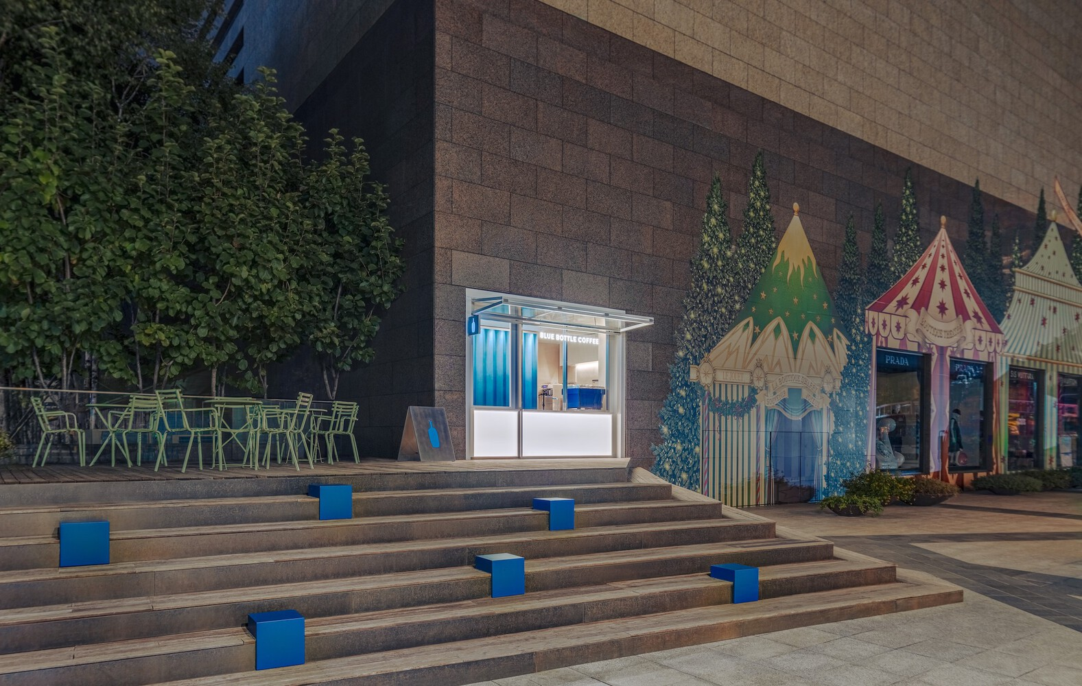 Illuminated kiosk between planted trees and decorative tent structures at evening