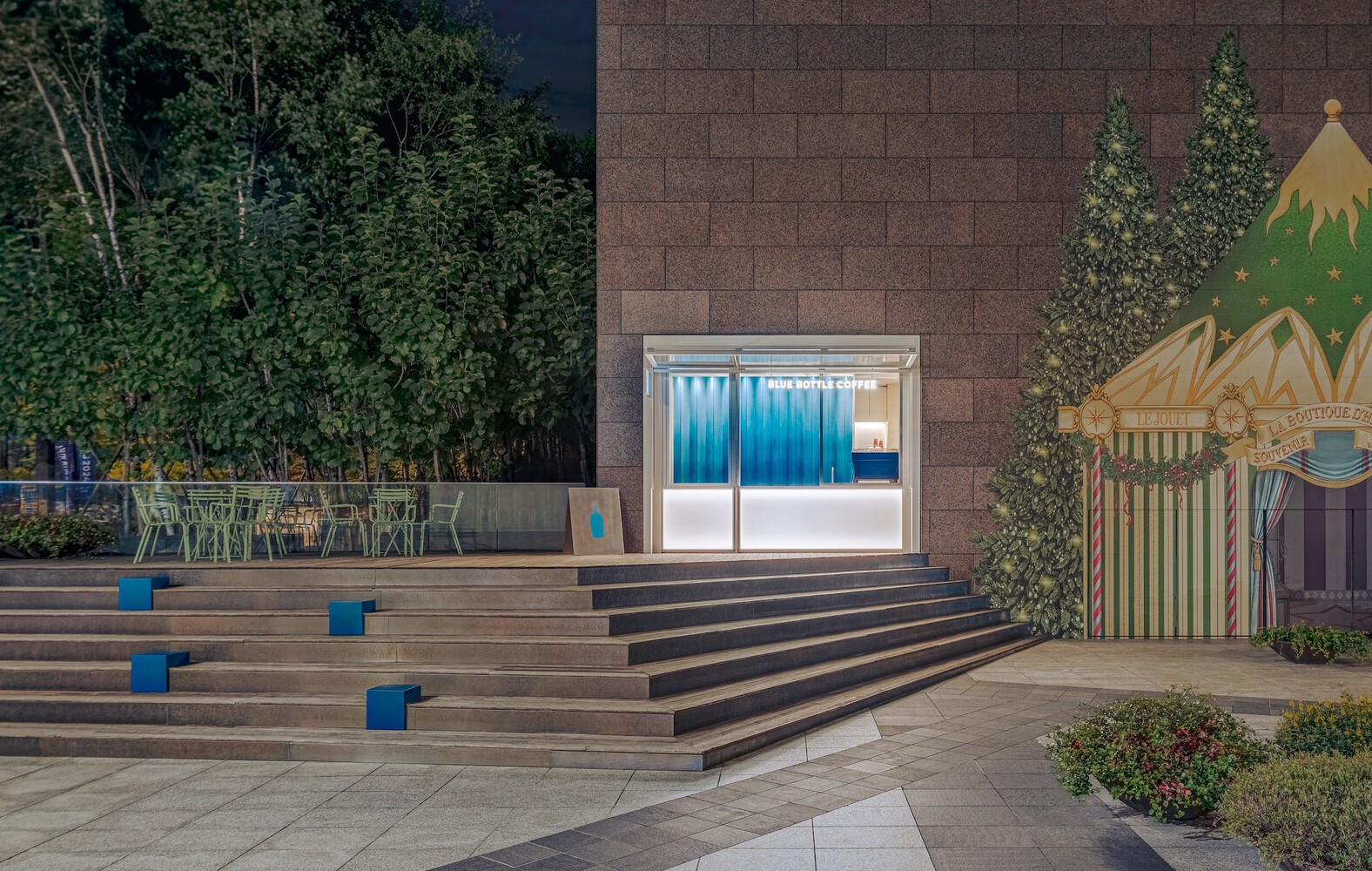 Timber stepped plaza with blue accent cubes leading to a glazed kiosk at night