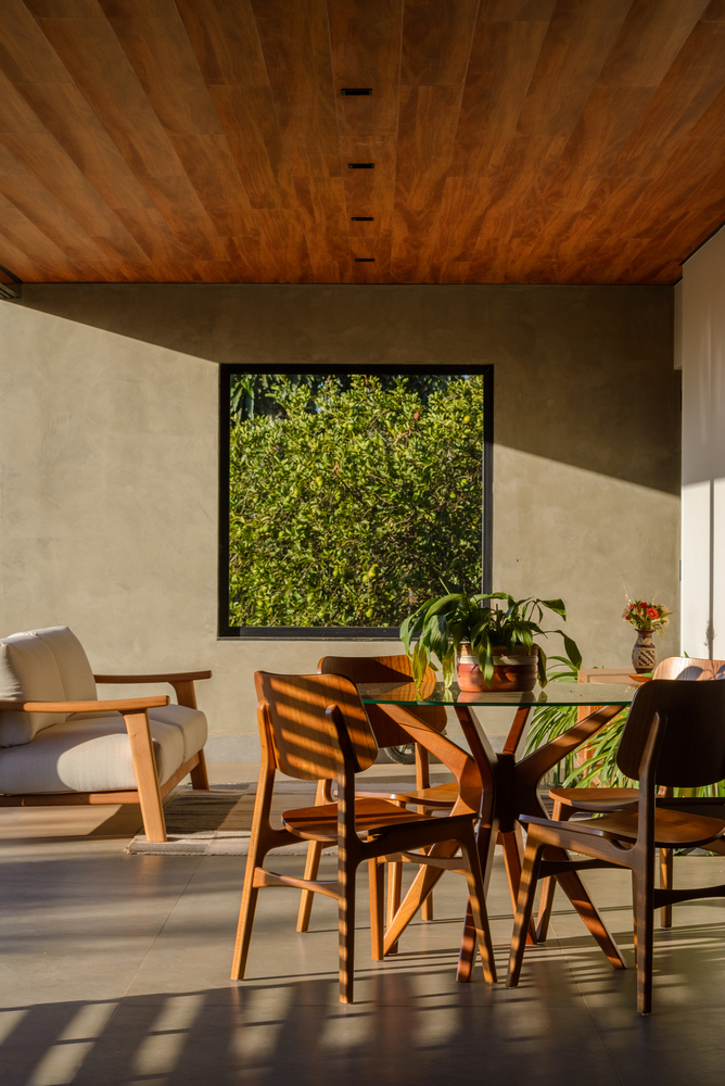 Dining area with timber ceiling, framed garden view, and sunlight casting shadows across the concrete floor