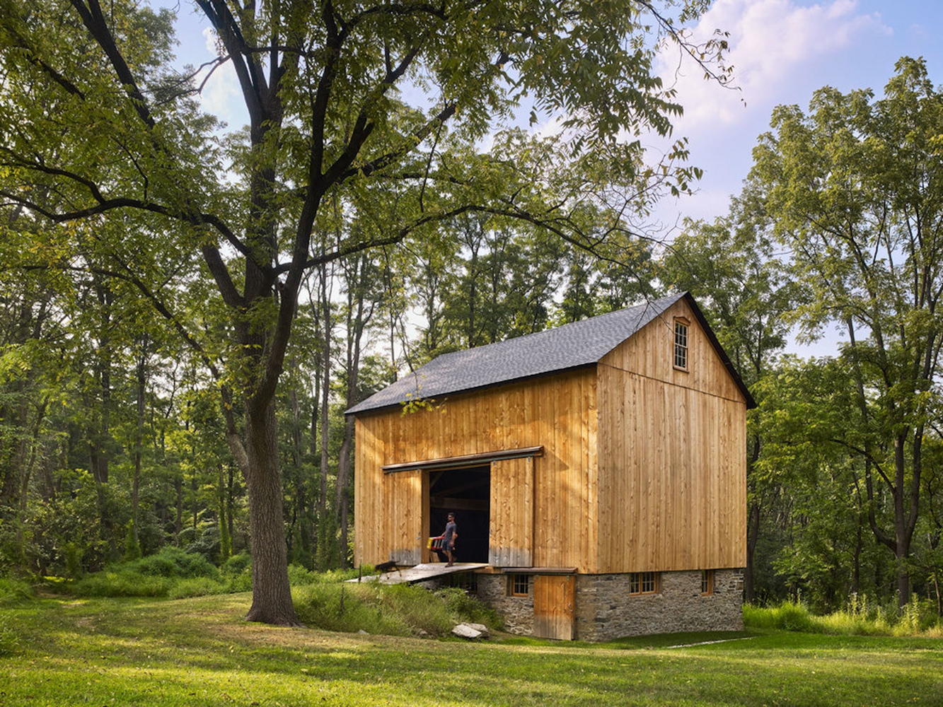 Timber-clad barn with stone base and large opening framed by mature deciduous trees on a clearing