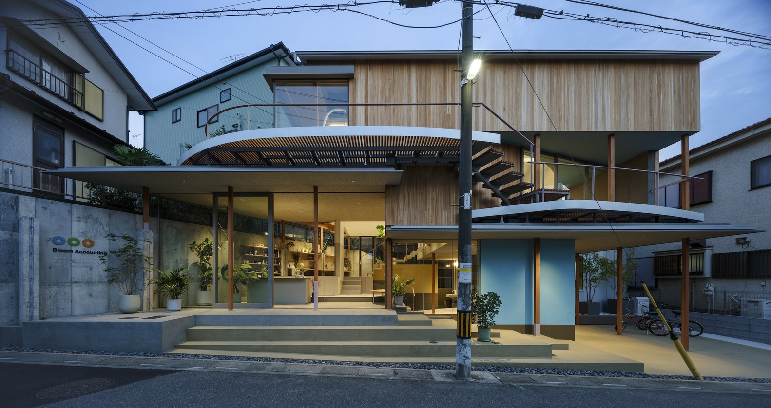 Street view at dusk showing timber-clad volumes and glazed ground floor with illuminated interiors and overhead wires
