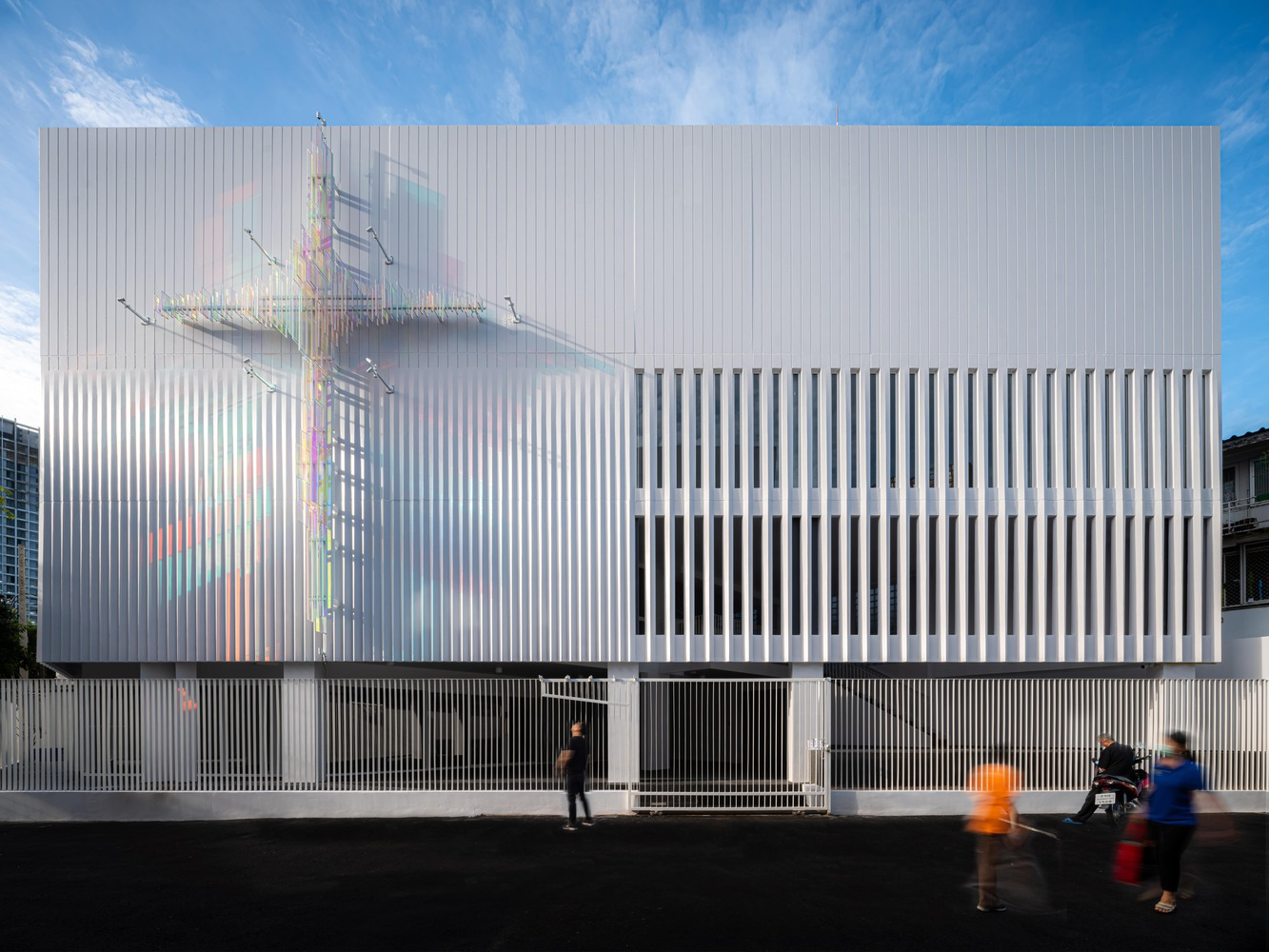 Street view of the white vertical-ribbed facade with dichroic glass cross sculpture and pedestrians passing by