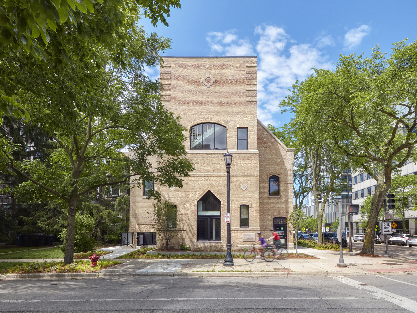 Street view of the buff brick facade with arched windows and a cyclist passing under leafy trees