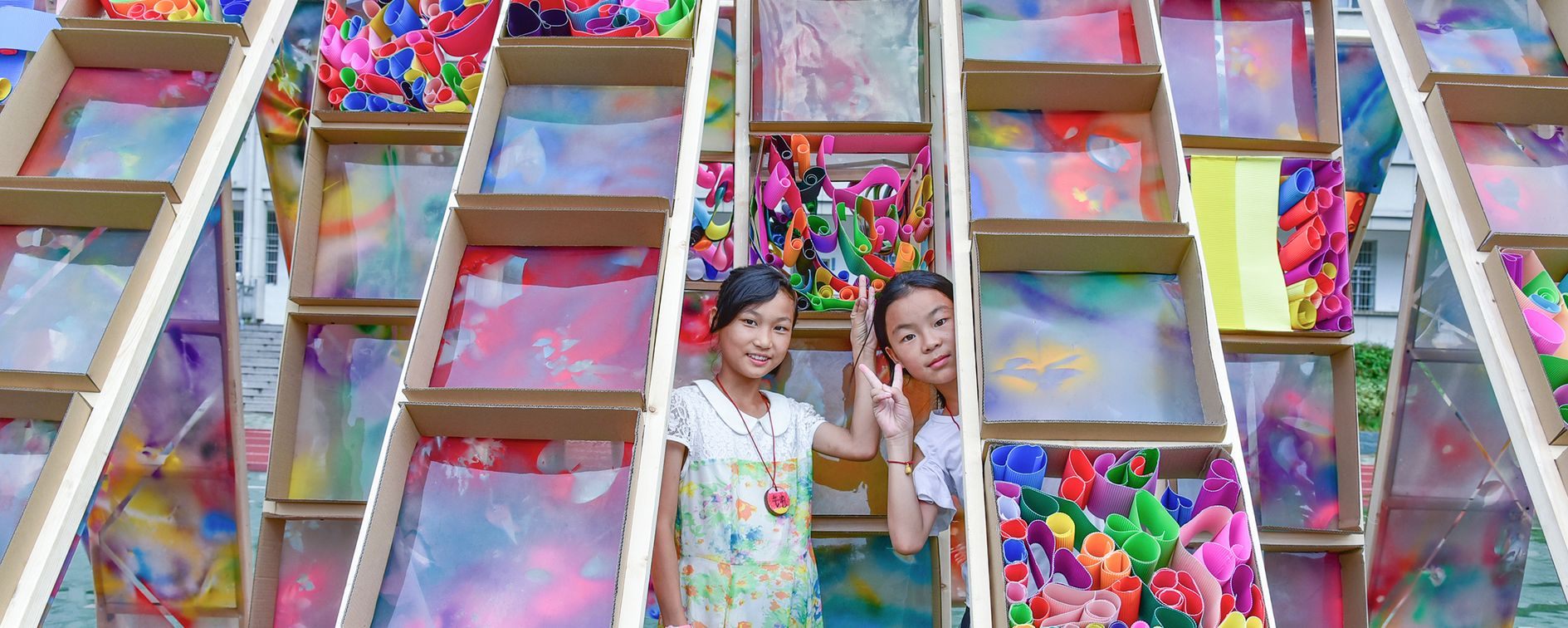 Two children pose beside the vibrant, hand-crafted wooden ladders of the Hi Ladders High pavilion, showcasing their creativity and design skills.