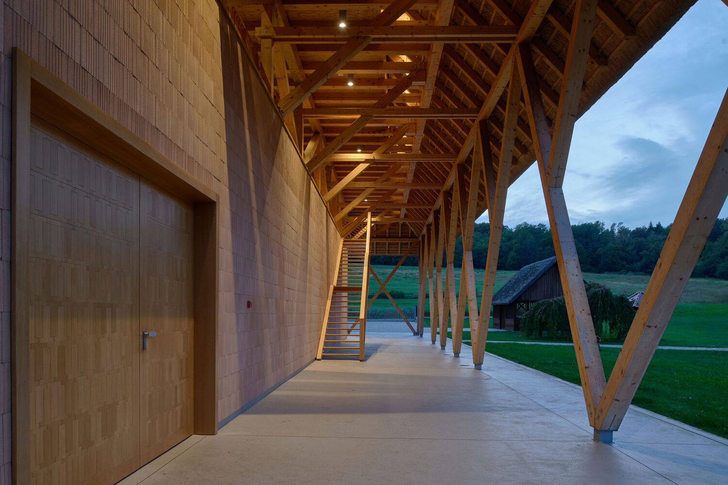 Covered walkway with exposed timber beams and diagonal columns along a tile wall at dusk