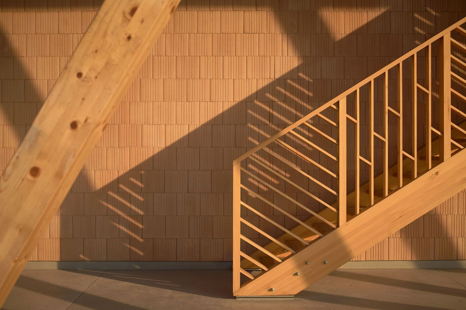 Timber staircase with diagonal railings casting shadows on a brick tile floor in warm sunlight