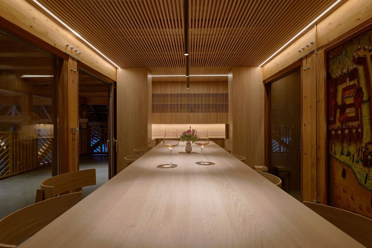 Dining hall with slatted timber ceiling, linear lighting, and a long pale wood table at night