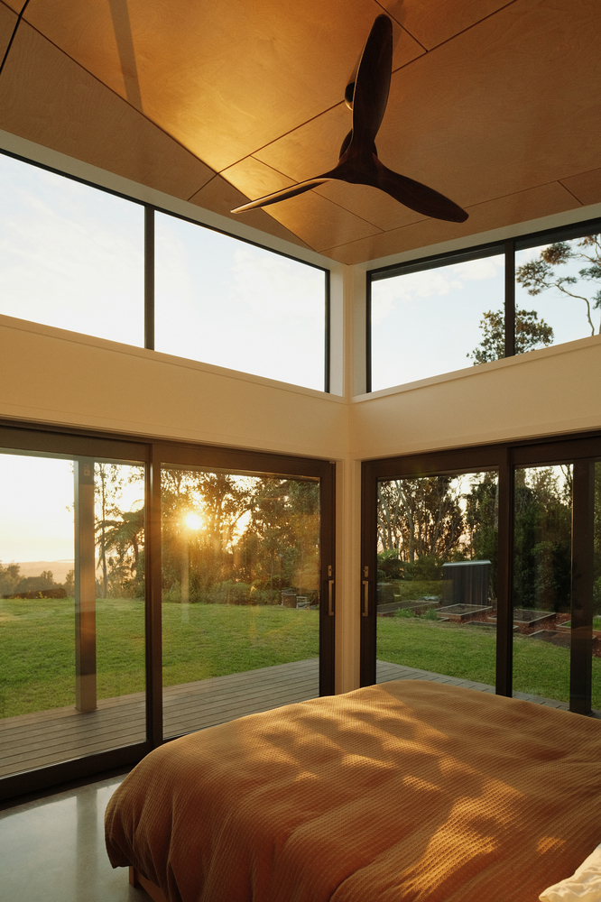 Interior bedroom corner with plywood ceiling panels and full-height glazing overlooking lawn at sunset