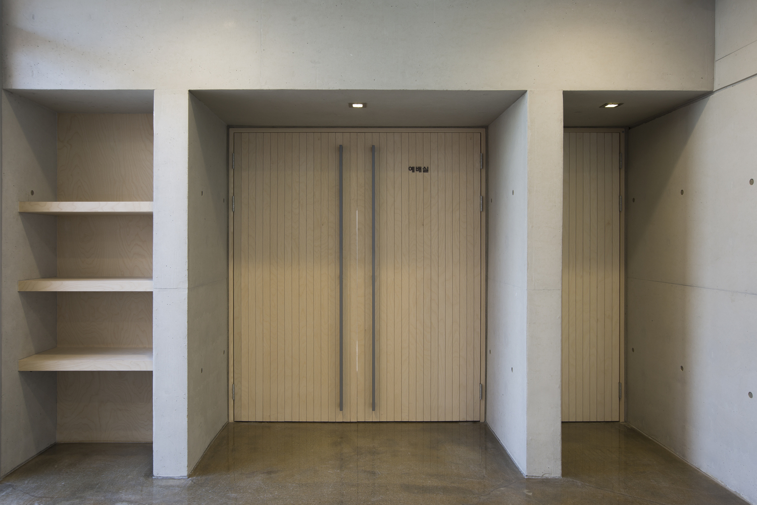 Entrance alcove with vertical wood door flanked by concrete walls and recessed shelving niche